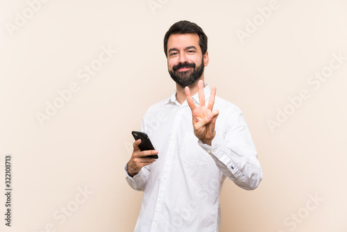 Fotografie, Tablou Young man with beard holding a mobile happy and counting three with fingers