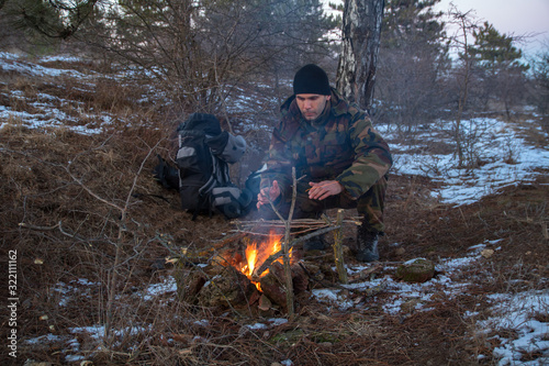 caucasian man in the camouflage sitting near the campfire in the winter forest, at night time