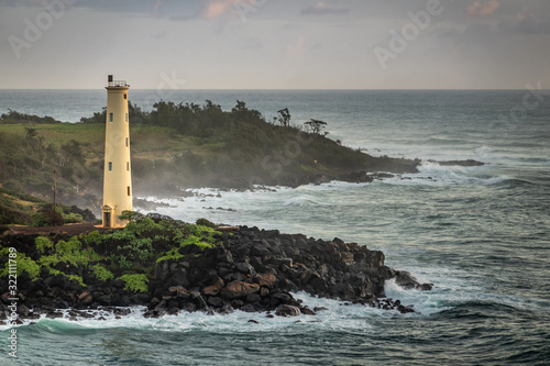 Nawiliwili, Kauai, Hawaii, USA. - January 16, 2020: Yellow Ninini lighthouse on black ocean-shore rocks early morning. Dark gray ocean under light gray sky. Some green foliage on land.