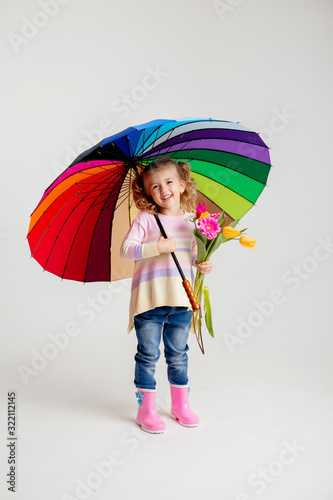 little blonde girl is smiling, standing in rubber boots holding spring flowers and a multi-colored umbrella on a white background