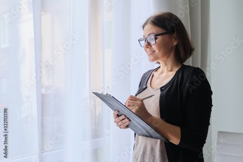 Mature woman teacher taking notes, standing near window in office photo