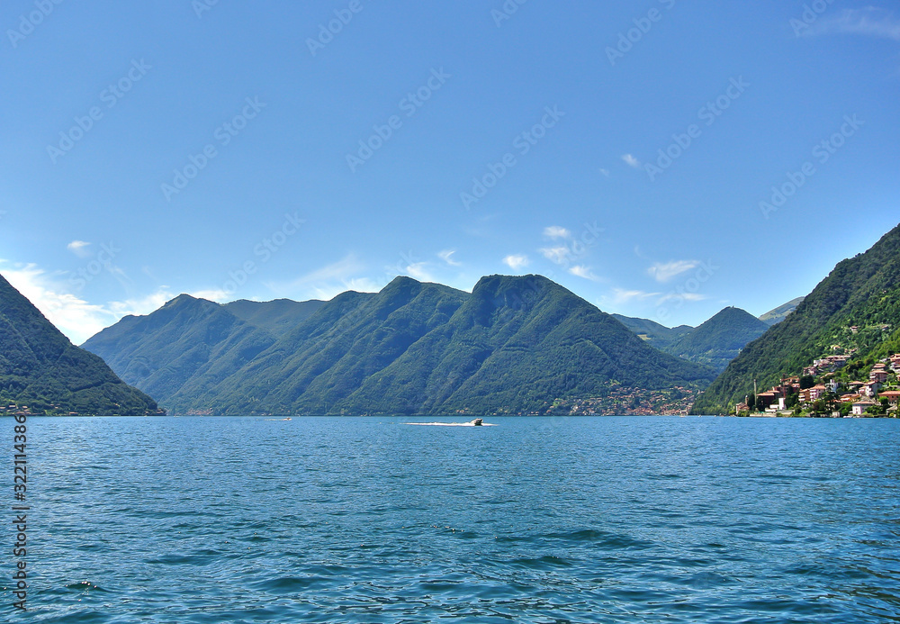 Lake Como in the Lombardy region (Lombardia) of Italy, green alpine mountains covered with trees, clouds and a small town in the distant, summer afternoon.