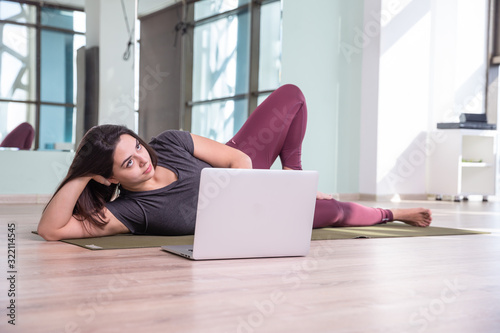 Photo of young woman practicing yoga with laptop indoor. Beautiful girl practice yoga in class. Yoga studio instructor. Blurred background.