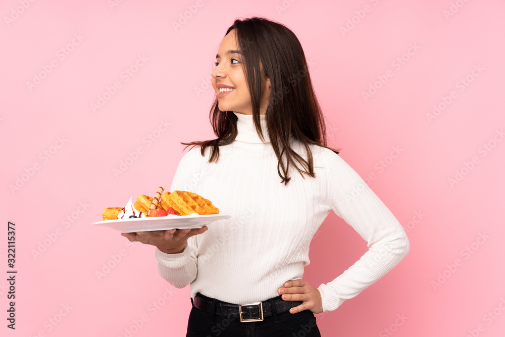 Young brunette woman holding waffles over isolated pink background looking side