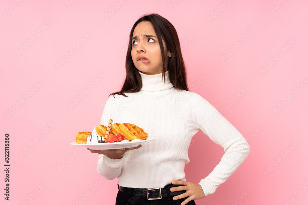Young brunette woman holding waffles over isolated pink background with confuse face expression Young brunette woman holding waffles over isolated pink background with confuse face expression