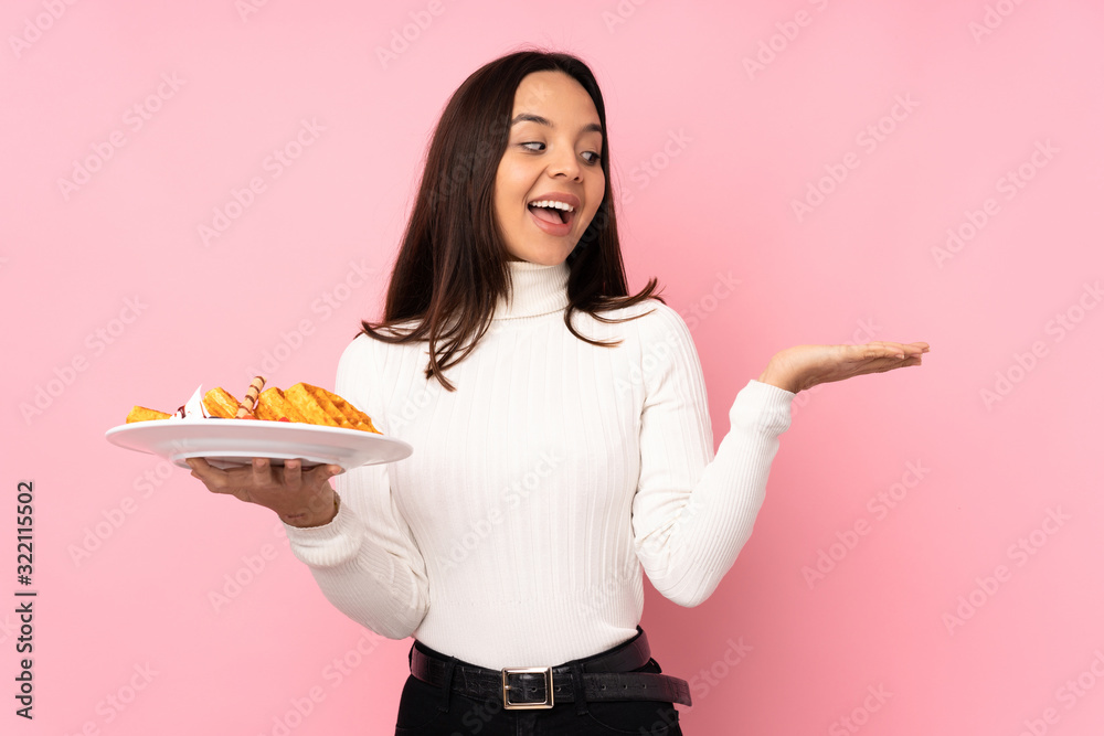 Young brunette woman holding waffles over isolated pink background holding copyspace with two hands