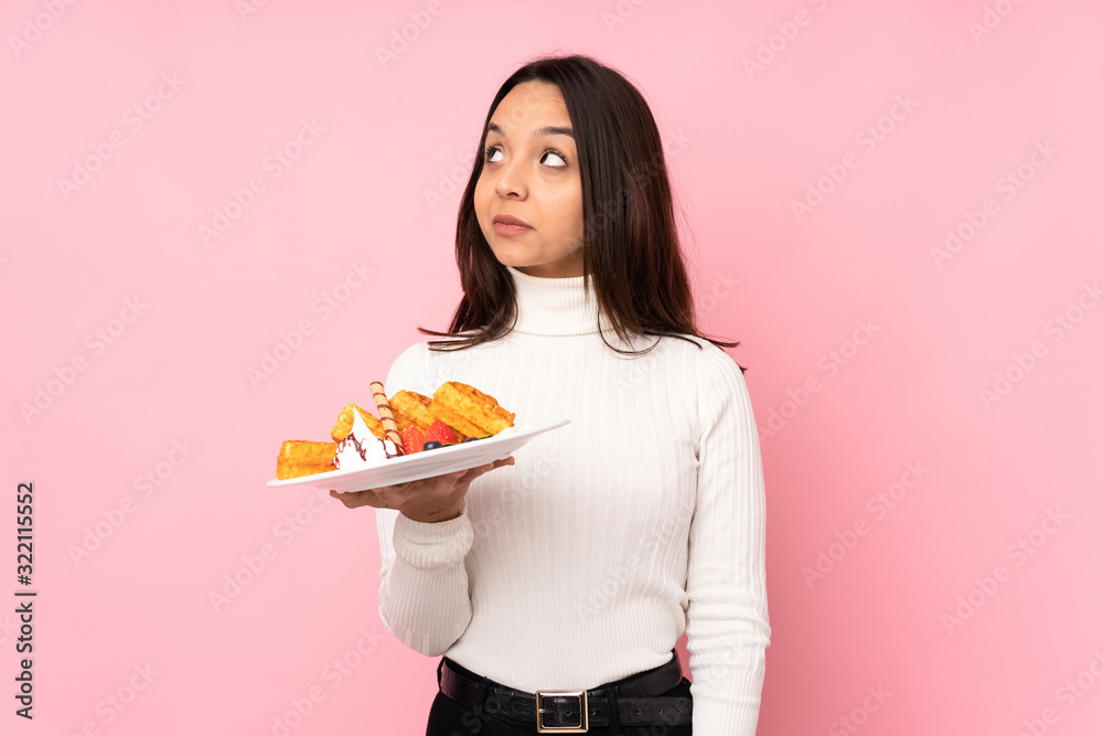 Young brunette woman holding waffles over isolated pink background making doubts gesture looking side Young brunette woman holding waffles over isolated pink background making doubts gesture looking side