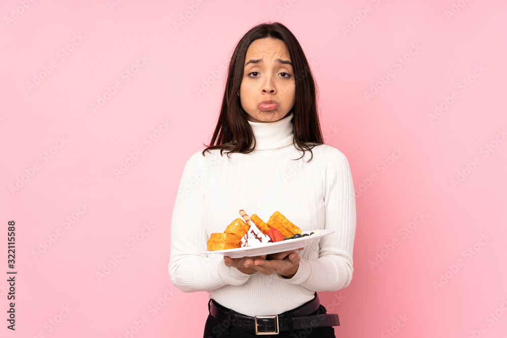 Young brunette woman holding waffles over isolated pink background keeps palm together. Person asks for something Young brunette woman holding waffles over isolated pink background keeps palm together. Person asks for something