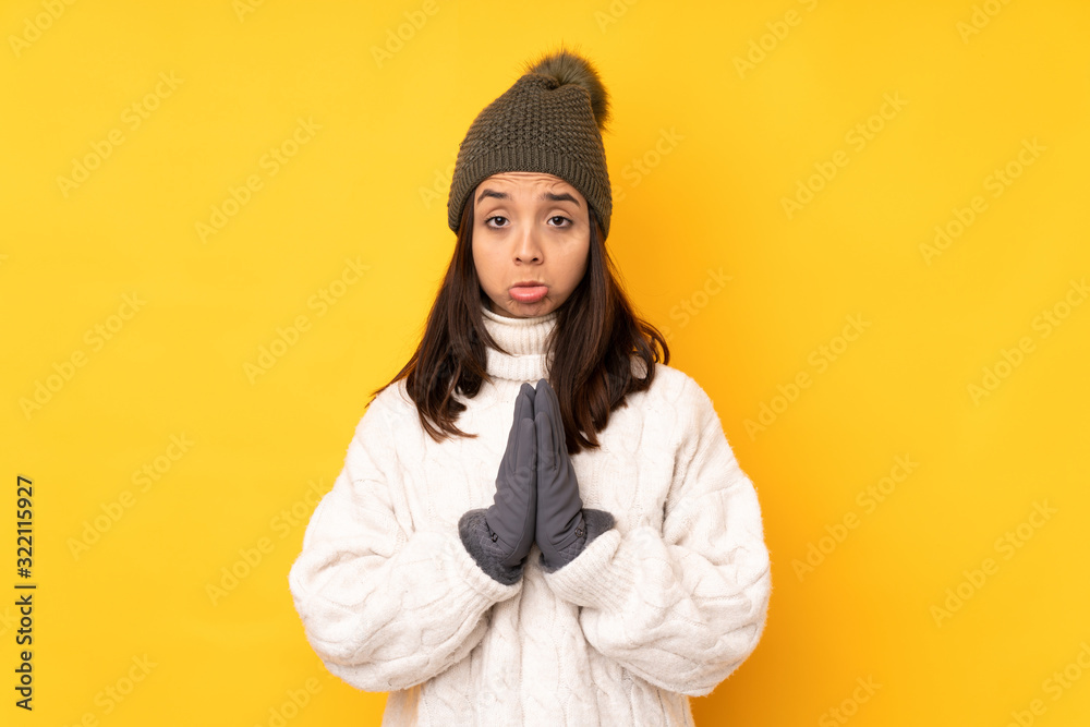 Young woman with winter hat over isolated yellow background keeps palm together. Person asks for something Young woman with winter hat over isolated yellow background keeps palm together. Person asks for something
