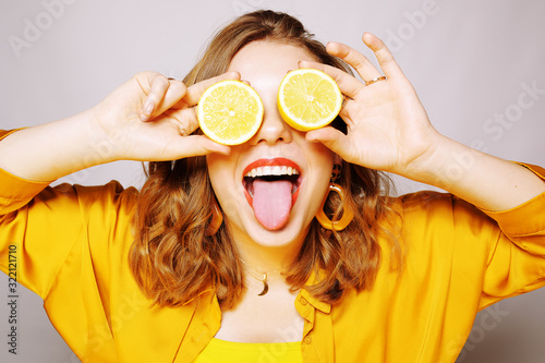 Portrait of a girl with a lemon. A girl in a bright yellow dress, with yellow earrings, holds a lemon. covering eye with lemon on a white background. Girl shows tongue, has fun.