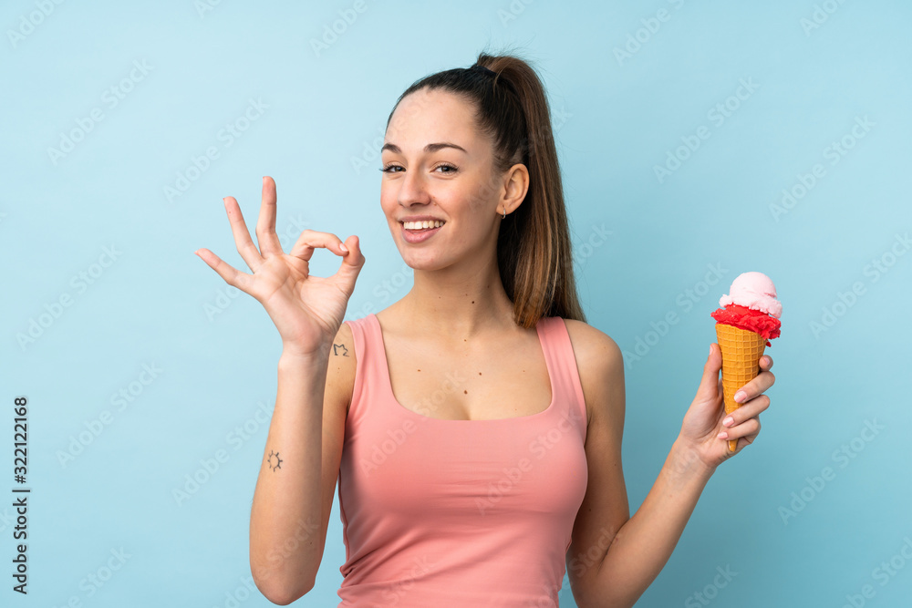 Young brunette woman with a cornet ice cream over isolated blue background showing ok sign with fingers Young brunette woman with a cornet ice cream over isolated blue background showing ok sign with fingers