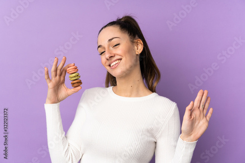 Young brunette woman over isolated purple background holding colorful French macarons and saluting