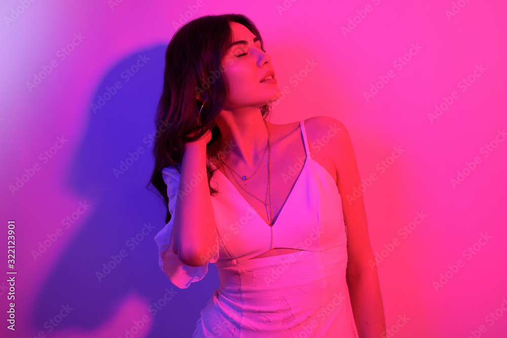 Brunette with long curled hair dancing in neon. A girl with professional makeup in red and blue light posing. Sexy model in a white dress at a party. Brunette with long curled hair dancing in neon. A girl with professional makeup in red and blue light posing. Sexy model in a white dress at a party.