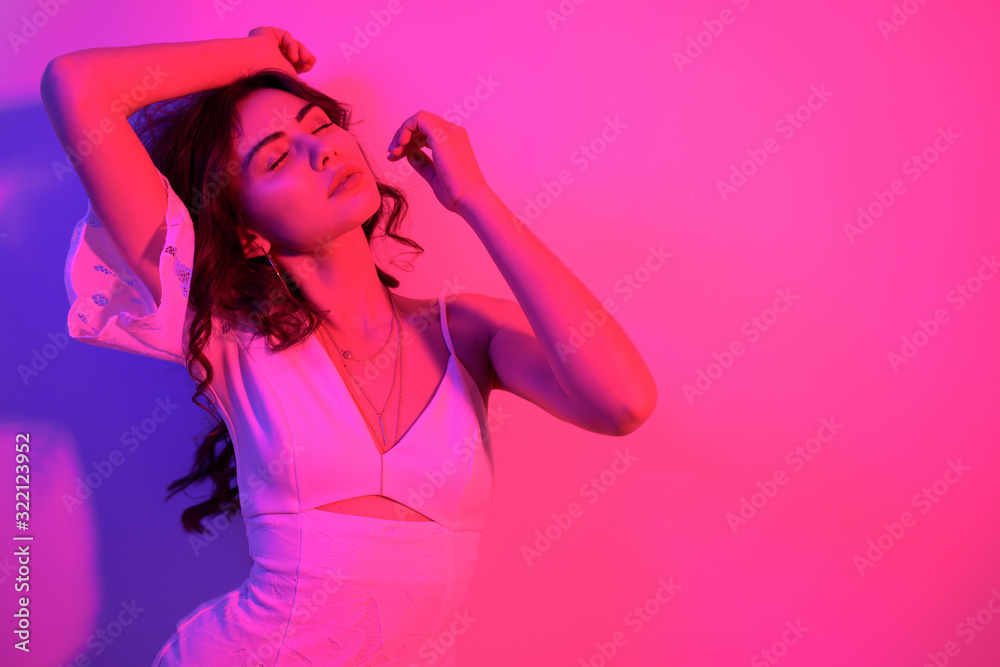 Brunette with long curled hair dancing in neon. A girl with professional makeup in red and blue light posing. Sexy model in a white dress at a party.