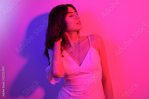 Brunette with long curled hair dancing in neon. A girl with professional makeup in red and blue light posing. Sexy model in a white dress at a party. © MoreThanProd