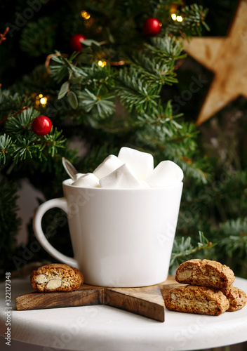 Coffee in a white cup with marshmallows. Morning festive coffee with traditional Italian cantuccini almond cookies. A cup of coffee on a background of green fir branches on a white stand.