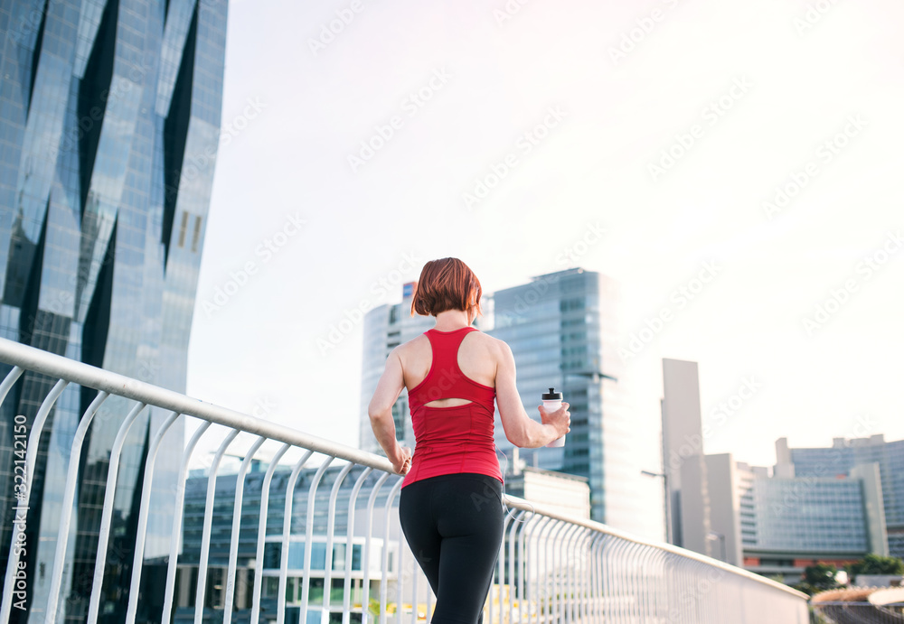 Rear view of young woman runner with water bottle jogging in city.
