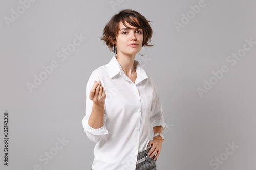 Confident young business woman in white shirt posing isolated on grey background. Achievement career wealth business concept. Mock up copy space. Rubbing fingers showing cash gesture asking for money.