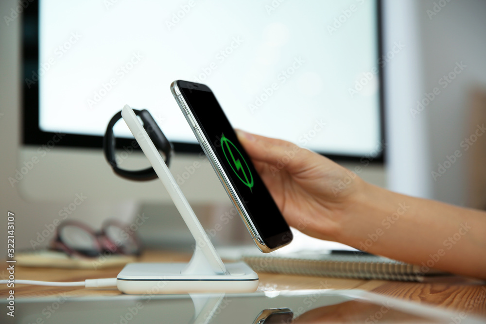 Fototapeta Woman putting mobile phone onto wireless charger at wooden table, closeup. Modern workplace accessory