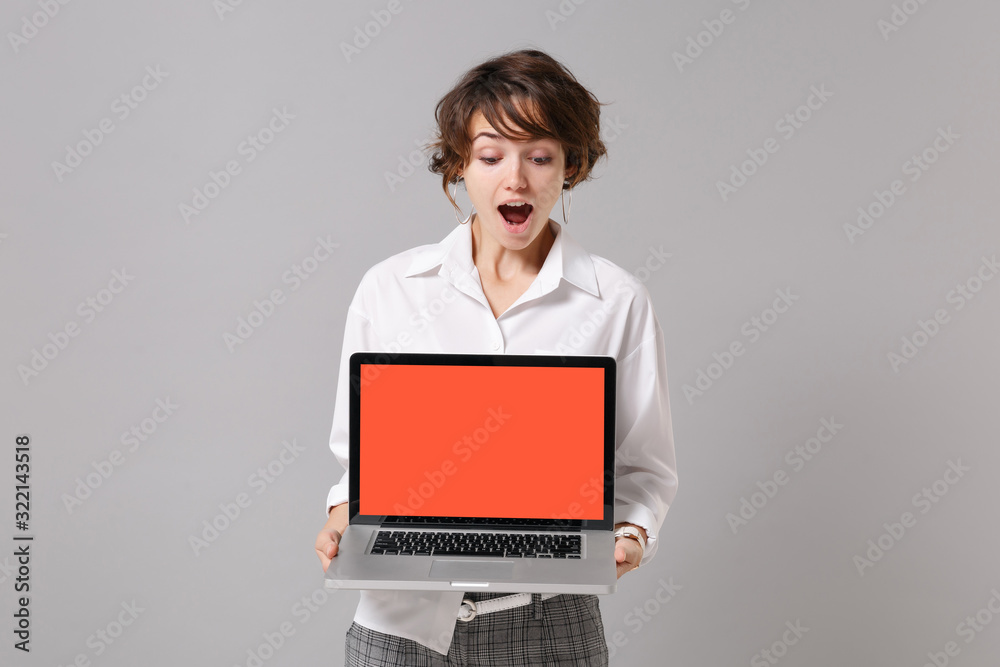 Shocked young business woman in white shirt posing isolated on grey wall background. Achievement career wealth business concept. Mock up copy space. Hold laptop pc computer with blank empty screen.