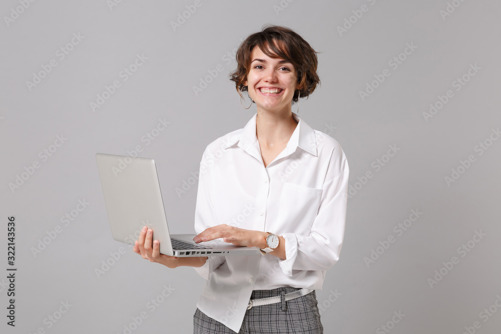 Cheerful young business woman in white shirt posing isolated on grey wall background studio portrait. Achievement career wealth business concept. Mock up copy space. Working on laptop pc computer. Cheerful young business woman in white shirt posing isolated on grey wall background studio portrait. Achievement career wealth business concept. Mock up copy space. Working on laptop pc computer.