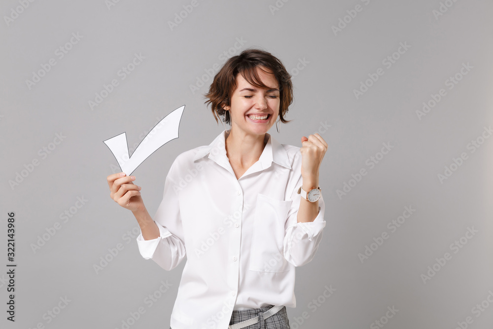 Fototapeta premium Happy young business woman in white shirt posing isolated on grey wall background in studio. Achievement career wealth business concept. Mock up copy space. Hold paper check mark doing winner gesture.