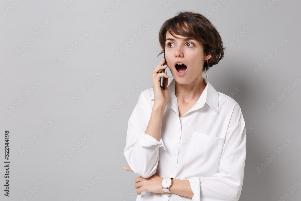 Shocked young business woman in white shirt posing isolated on grey background studio portrait. Achievement career wealth business concept. Mock up copy space. Talking on mobile phone, looking aside. Shocked young business woman in white shirt posing isolated on grey background studio portrait. Achievement career wealth business concept. Mock up copy space. Talking on mobile phone, looking aside.