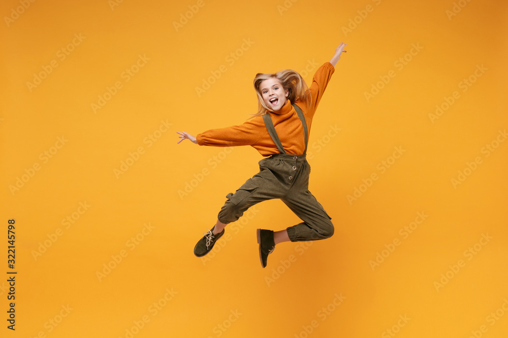 Cheerful little kid girl 12-13 years old in turtleneck, jumpsuit isolated on orange yellow wall background in studio. Childhood lifestyle concept. Mock up copy space. Jumping spreading hands and legs.