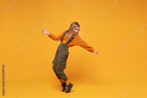 Funny little blonde kid girl 12-13 years old in turtleneck, jumpsuit isolated on orange yellow background. Childhood lifestyle concept. Mock up copy space. Dancing, standing on toes, spreading hands.