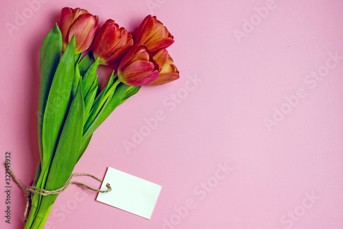 Fototapeta Naklejka Na Ścianę i Meble -  Bouquet of red tulips on a pink background with a tag for text.Bouquet of red tulips on a pink background with a tag for text. Top view,copy space. The concept of a greeting card for women's day