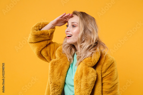 Beautiful young blonde woman girl in yellow fur coat posing isolated on orange background in studio. People lifestyle concept. Mock up copy space. Holding hand at forehead looking far away distance. © ViDi Studio