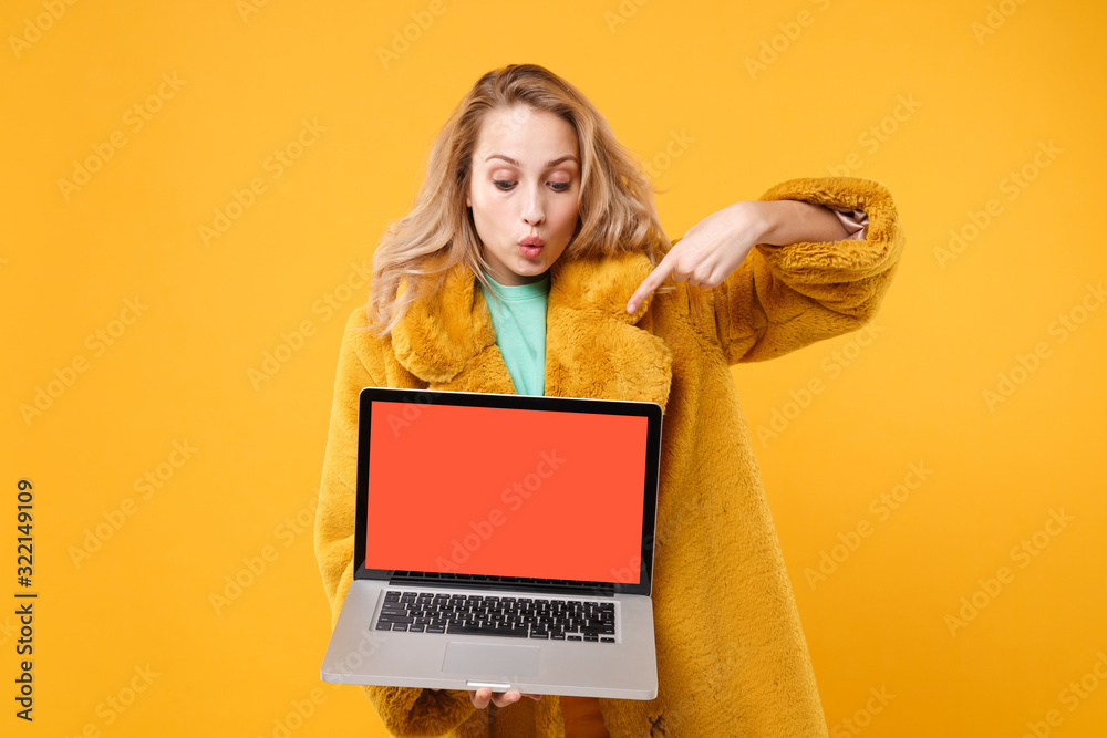 Naklejka premium Amazed young blonde woman in yellow fur coat isolated on orange background in studio. People lifestyle concept. Mock up copy space. Pointing index finger on laptop pc computer with blank empty screen.