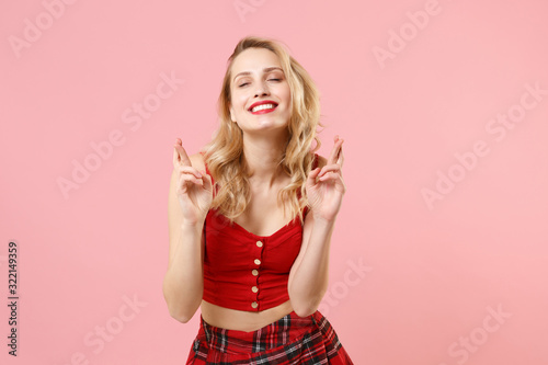 Smiling woman in red sexy clothes posing isolated on pastel pink background. People lifestyle concept. Mock up copy space. Waiting for special moment keeping fingers crossed, eyes closed, biting lips. © ViDi Studio