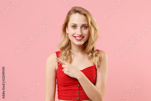 Smiling young blonde woman girl in red sexy clothes posing isolated on pastel pink wall background studio portrait. People emotions lifestyle concept. Mock up copy space. Pointing index finger aside.