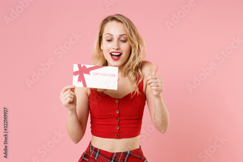 Cheerful young blonde woman girl in red sexy clothes isolated on pastel pink background studio portrait. People emotions lifestyle concept. Mock up copy space. Hold gift certificate, showing thumb up.