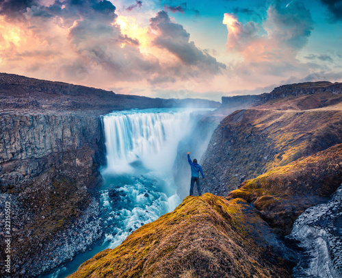 Tourist standing on the shore of falling water of the most powerful waterfall in Europe - Dettifoss. Impressive summer sunrise in Jokulsargljufur National Park, Iceland.