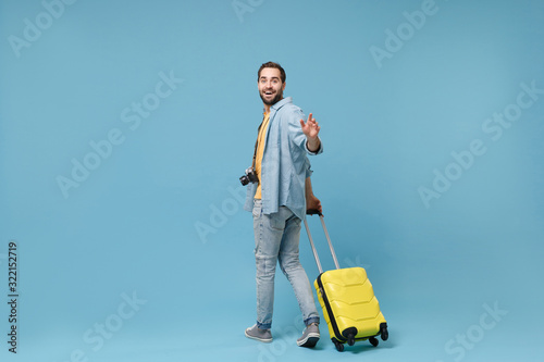 Back rear view of traveler tourist man with photo camera isolated on blue background. Passenger traveling abroad on weekends. Air flight journey. Holding suitcase, waving hand for greeting catch taxi.
