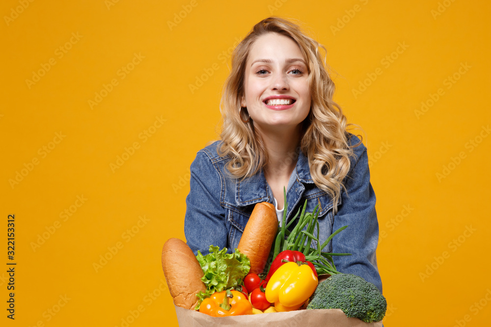 Smiling young woman in denim clothes posing isolated on yellow orange background. Delivery service from shop or restaurant concept. Hold brown craft paper bag for takeaway mock up with food products. Smiling young woman in denim clothes posing isolated on yellow orange background. Delivery service from shop or restaurant concept. Hold brown craft paper bag for takeaway mock up with food products.