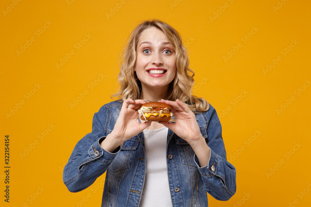 Smiling young woman in denim clothes isolated on yellow orange wall background studio portrait. Proper nutrition or American classic fast food concept. Mock up copy space. Holding in hands burger. Smiling young woman in denim clothes isolated on yellow orange wall background studio portrait. Proper nutrition or American classic fast food concept. Mock up copy space. Holding in hands burger.