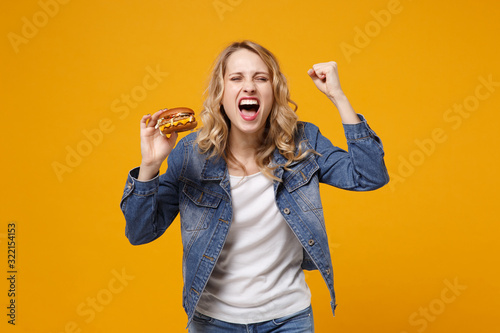 Crazy young woman in denim clothes isolated on yellow orange background. Proper nutrition or American classic fast food concept. Hold burger keeping eyes closed screaming clenching fist like winner.