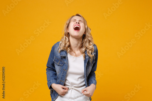 Screaming crying young woman in denim clothes isolated on orange background. Proper nutrition losing weight healthy lifestyle dieting concept. Measuring waist with measure tape keeping eyes closed.