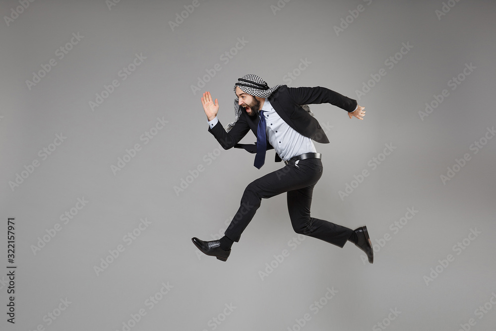 Cheerful young bearded arabian muslim businessman in keffiyeh kafiya ring igal agal classic black suit isolated on gray background. Achievement career wealth business concept. Jumping like running.