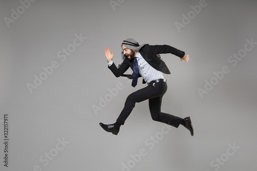 Cheerful young bearded arabian muslim businessman in keffiyeh kafiya ring igal agal classic black suit isolated on gray background. Achievement career wealth business concept. Jumping like running.