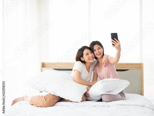 Asian young two girl smile and cuddle together for selfie photo and snap by smartphone technology device with best friend lifestyle teen leisure on white bed and pillows in bedroom at minimal home