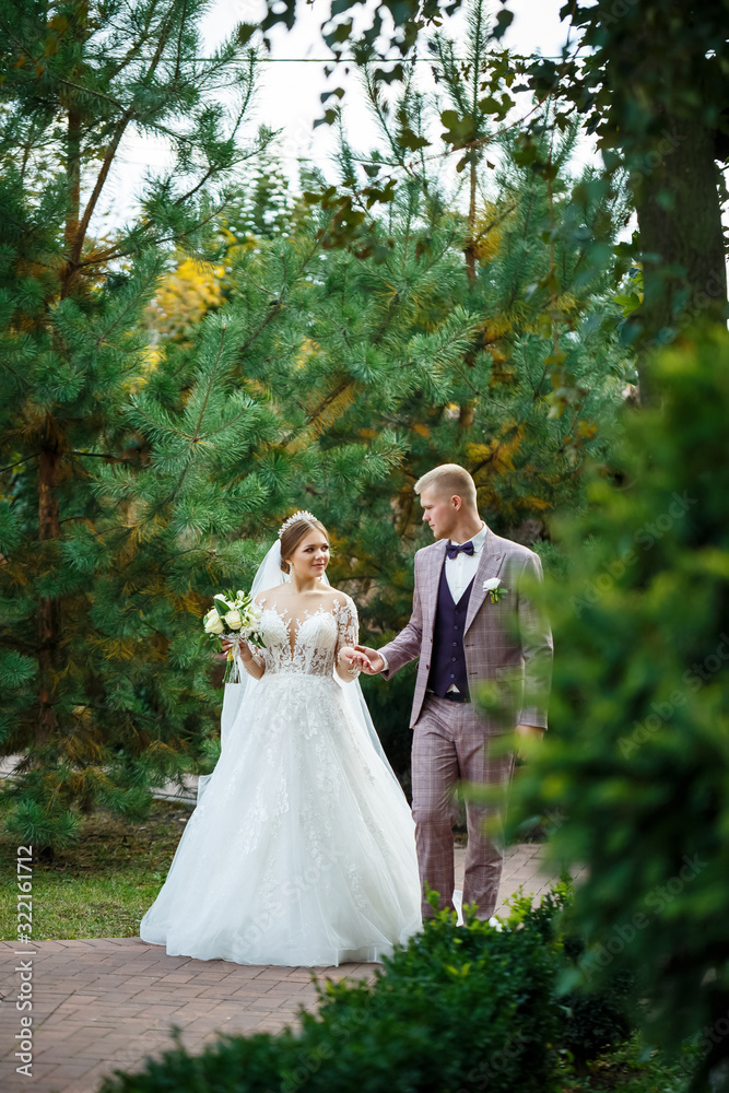Bride in white dress and groom in costume cuddle and walk in the park