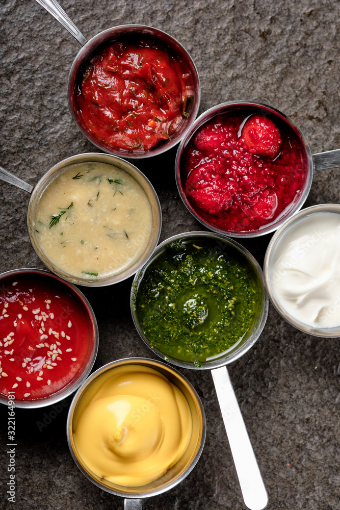 Set of different sauces - ketchup, mayonnaise, barbecue, mustard, grain hills, pesto, adzhika, chutney, tkemali sauce on on black stone background. Top view copy space