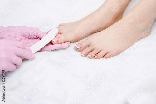 Specialist in beauty salon making french pedicure for client. Closeup young well-groomed female foots. Manicurist doing professional pedicure. Master files nails to client.