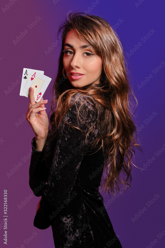 Brunette girl in black velvet dress showing two playing cards, posing sideways on coloful background. Gambling entertainment, poker, casino. Close-up. Brunette girl in black velvet dress showing two playing cards, posing sideways on coloful background. Gambling entertainment, poker, casino. Close-up.