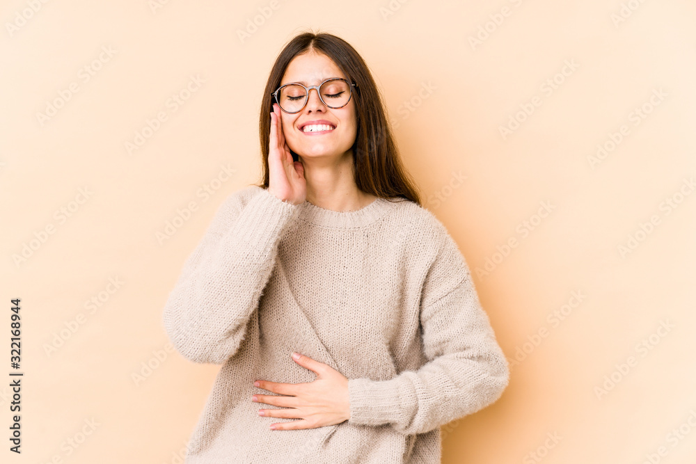 Young caucasian woman isolated on beige background laughs happily and has fun keeping hands on stomach. Young caucasian woman isolated on beige background laughs happily and has fun keeping hands on stomach.