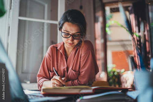 Young concentrated woman in spectacles writing notes in notebook while working in modern office, texting business message communicating online with client feels satisfied successful concept photo